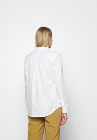 Chemise blanche à manches longues avec col, présentant une couture droite à l'arrière et des poignets à boutons, associée à un pantalon jaune moutarde.