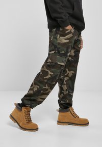 Camo cargo pants in green and brown with side pockets, paired with tan lace-up boots featuring a textured surface and rubber soles.