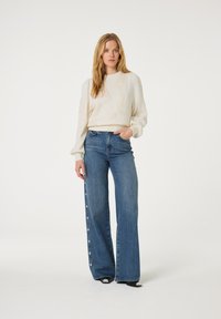 Witte gebreide trui met textuur van bladeren, gedragen met hooggerande, wijde blauwe jeans met hartvormige knopen aan de zijkant.