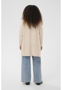 Ilgas garbanotas plaukas turintis žmogus dėvi bežinį megztinį, plačius šviesiai mėlynus džinsus ir juodus batus, stovi prieš paprastą foną.