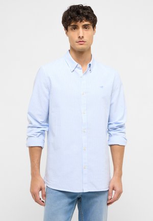 Junger Mann mit lockigem dunklem Haar, der ein hellblau-weiß vertikal gestreiftes Hemd mit hochgekrempelten Ärmeln und hellblaue Jeans trägt.