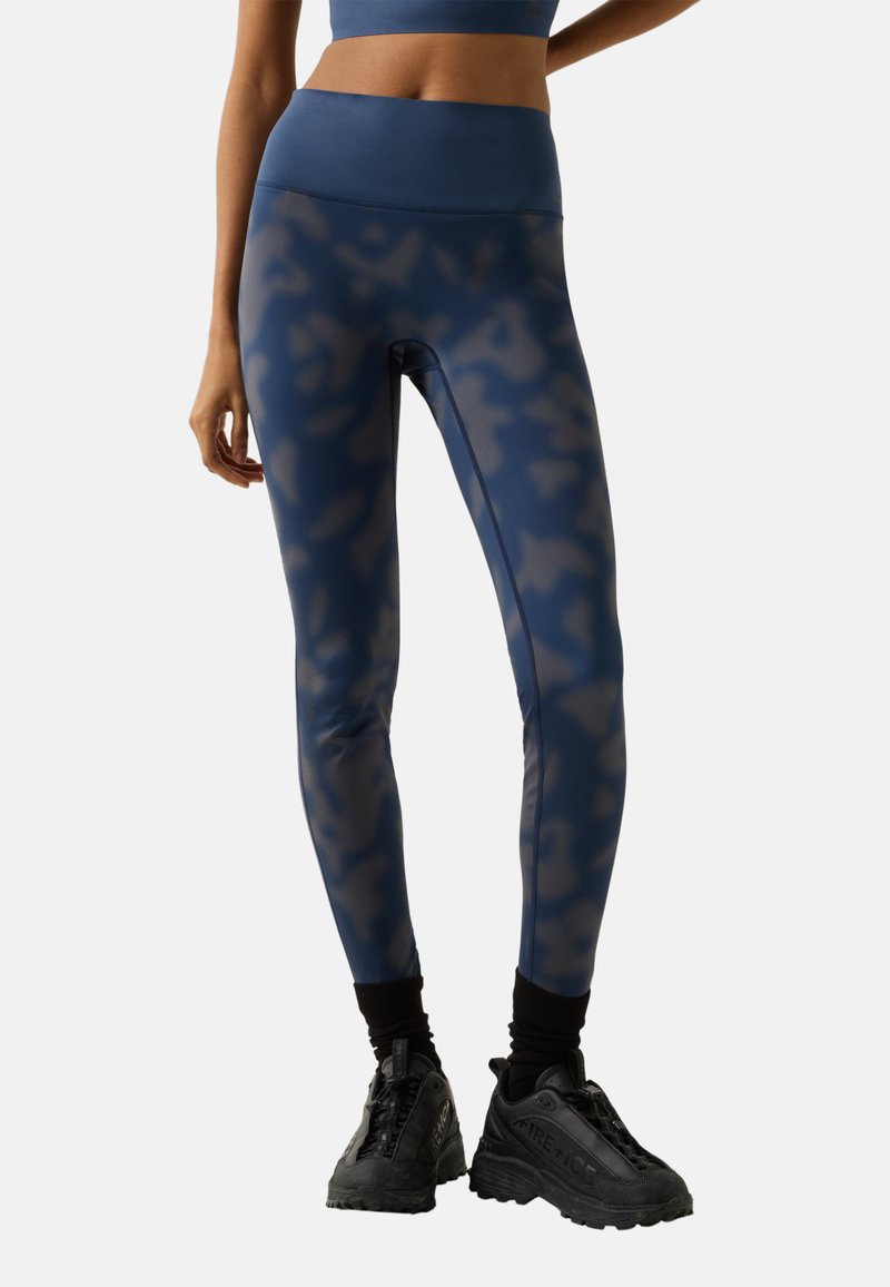 Kvinde iført blå leggings med camouflage-mønster, sorte sokker og sorte chunky sneakers, stående mod en ensfarvet baggrund.
