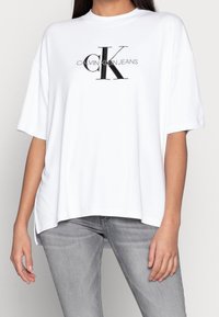 White cotton T-shirt with short sleeves, loose fit, featuring a black "CK" logo and "CALVIN KLEIN JEANS" text in a clean font.