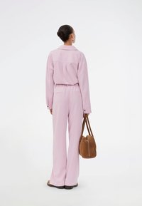 Φωτεινό ροζ, υπερμεγέθη πουκάμισο με κουμπιά και λεπτομέρειες στην πλάτη, συνδυασμένο με αντίστοιχες φαρδιές παντελόνες. Το άτομο κρατά μια καφέ τσάντα χειρός.