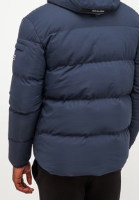 Tmavě modrá péřová bunda s texturovaným povrchem, vybavená kapucí, vodorovnými švy a malým logem na rukávu.