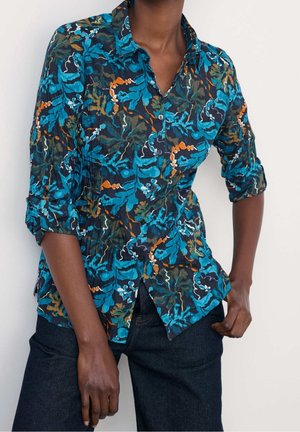 Personne portant une chemise boutonnée à manches longues avec des motifs abstraits de feuilles bleues, orange et vertes et un jean foncé sur un fond uni.
