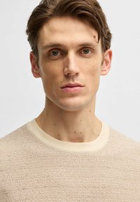 Maglione a maglia beige con texture, colletto a girocollo color crema, con un motivo sottile e una finitura morbida, indossato da un modello con i capelli corti.