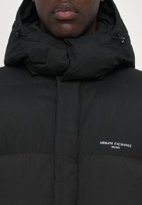 Čierna nap puffrová bunda s vysokým golierom a kapucňou. Obsahuje jemné prešitie a biele logo "ARMANI EXCHANGE MILANO" na ľavej strane.