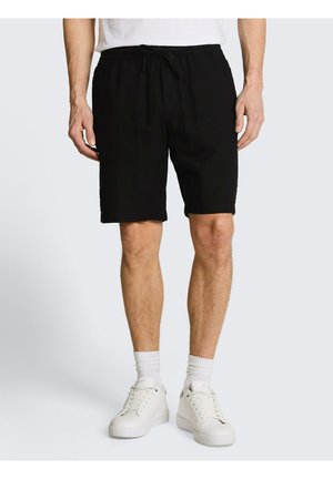 Męski model noszący czarne szorty ze ściągaczem, białe skarpetki i białe sneakersy, stojący na tle jasnoszarego tła.