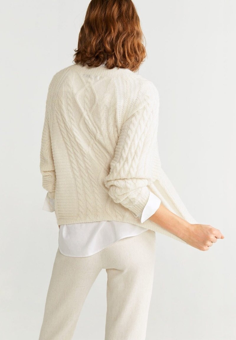 Femme aux cheveux mi-longs, portant un pull crème en maille torsadée et un pantalon assorti, tenant l'ourlet du pull, tournée de dos sur un fond uni.