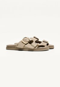 Bruna mockasin sandaler i mocka med två justerbara guldbuckles, en texturerad gummisula och öppen tådesign.