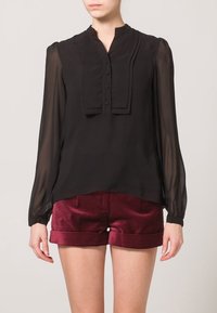 Schwarze transparente Bluse mit hohem Kragen, plissiertem Vorderteil und Knopfdetails. Lange Ärmel mit enganliegenden Manschetten, kombiniert mit burgunderfarbenen Shorts.