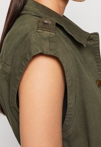 Veste sans manches vert olive avec un col, présentant des détails de boutons sur les épaules et des accents de couture. Le matériau semble être du coton ou un tissu similaire.