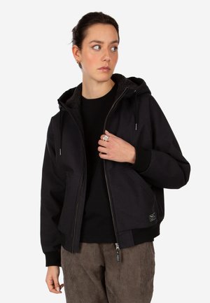 Frau mit dunklen, zurückgebundenen Haaren trägt eine schwarze Kapuzenjacke über einem schwarzen Shirt und braune Kordhosen und schaut nach links.