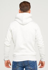 Mann mit braunen Haaren, der einen hellgrauen Kapuzenpullover und blaue, abgenutzte Jeans trägt, von hinten vor einem schlichten Hintergrund gezeigt.