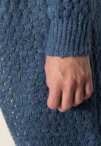 Prenda de punto azul con un patrón suelto y calado, puños acanalados y una textura suave, mostrando una mano descansando sobre la tela.