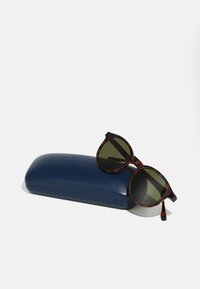 Gafas de sol redondas de carey con lentes verdes, descansando sobre un estuche texturizado de color azul oscuro grabado con "Lacoste".