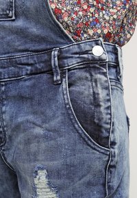 Salopette en denim bleu délavé avec un motif usé. Comprend un bouton en métal, des poches et un tissu à motif floral à l'intérieur des bretelles.
