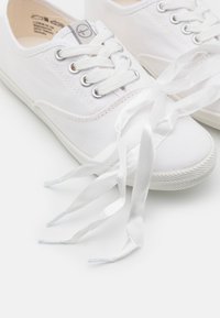 Baskets en toile blanche avec bout rond, œillets argentés et lacets plats blancs. Semelle en caoutchouc texturée et petit logo sur la languette.