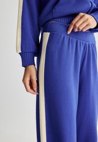 Pantaloni de jogging din polar albastri, cu o dunga pe side de culoare crem, brâu elastic și manșete cu țesătură cu nervuri. Textura moale și croiala lejeră sunt accentuate.