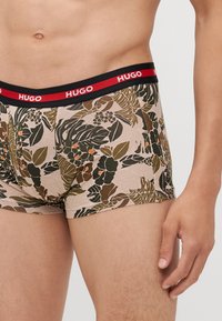 Le short de boxeur présente un motif floral dans des tons terreux, avec des feuilles vertes et des accents orange. La ceinture est noire avec l'inscription "HUGO" en rouge.