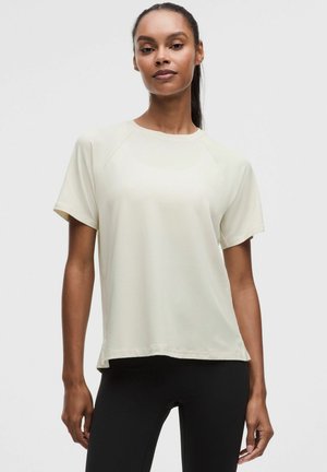 Femme aux cheveux attachés, portant un t-shirt crème uni à manches courtes et des leggings noirs sur un fond gris clair.