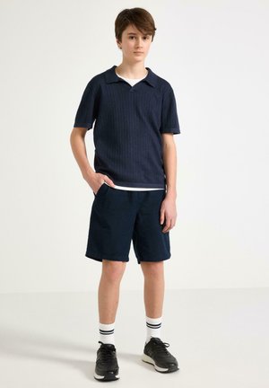 Teini-ikäinen poika seisoo kädet taskuissa, päällään laivastonsininen lyhythihainen poolopaita, tummat shortsit, valkoiset raidalliset sukat ja mustat lenkkarit valkoisella taustalla.