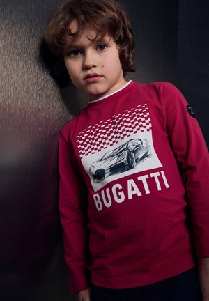 Ragazzo giovane con capelli castano medio che indossa una maglia rossa a maniche lunghe con uno schizzo di un'auto Bugatti e un motivo a scacchi.