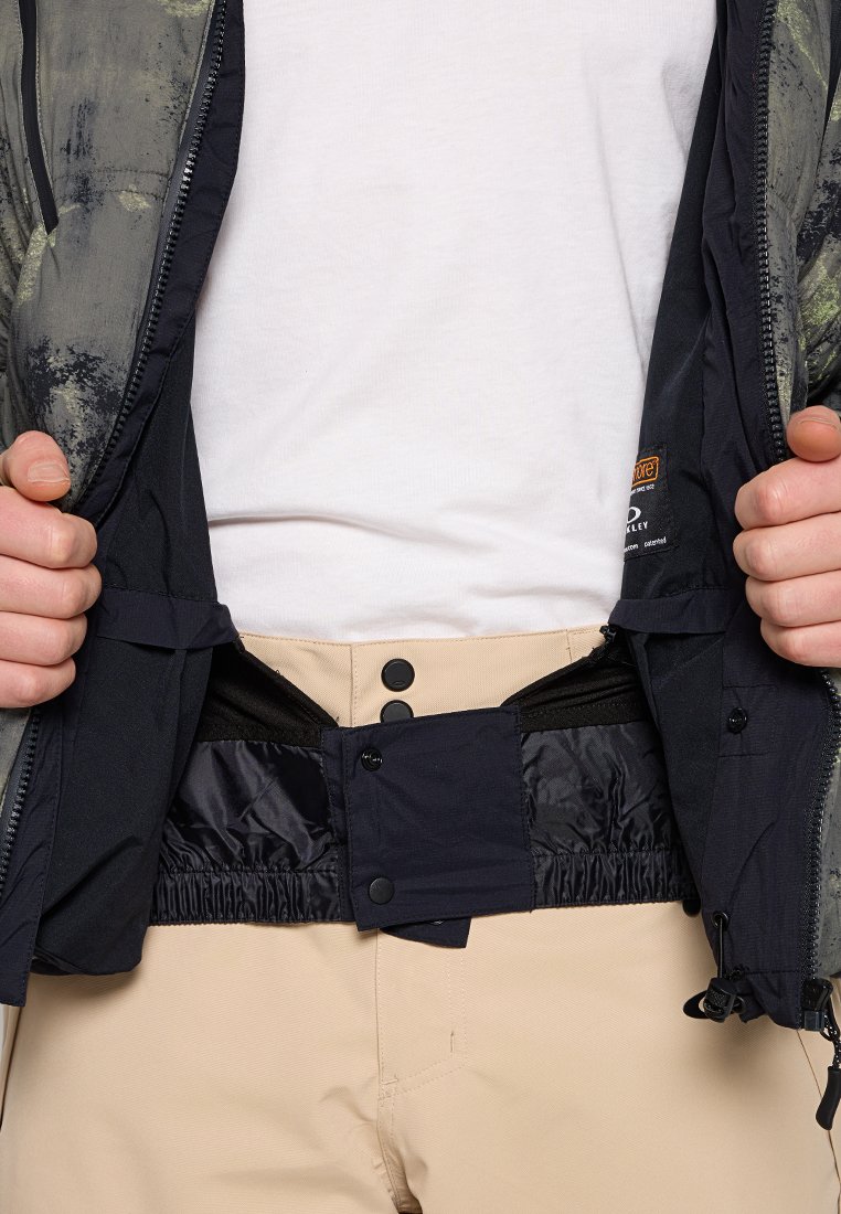 Des mains tenant une veste camouflage ouverte pour révéler un pantalon beige avec une taille ajustable noire et des boutons-pression.