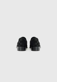 Chaussures en daim noir avec une texture lisse, présentant un motif de logo et un petit talon noir large. La vue arrière met en avant des lignes épurées et un design minimal.