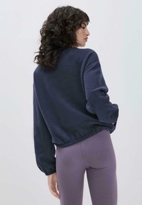 Marineblaues Fleece-Sweatshirt mit lockerer Passform, elastischem Saum und gerafften Ärmeln. Getragen mit lila Leggings; glatte Textur, lässiges Design.