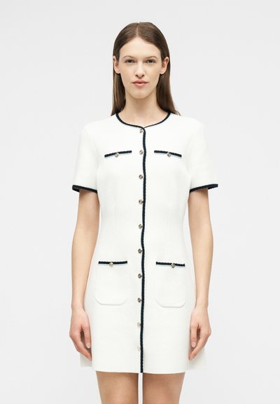 Γυναίκα που φοράει λευκό φόρεμα με κοντά μανίκια, κουμπωτό, με μαύρη επένδυση και τέσσερις τσέπες μπροστά, μπροστά σε απλό ανοιχτό φόντο.