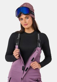 Skikleding met een paarse geïsoleerde jumpsuit met zwarte accenten, een ribbelige mauve muts en blauw-getinte skibril. Rits- en klittenbanddetails zijn zichtbaar.