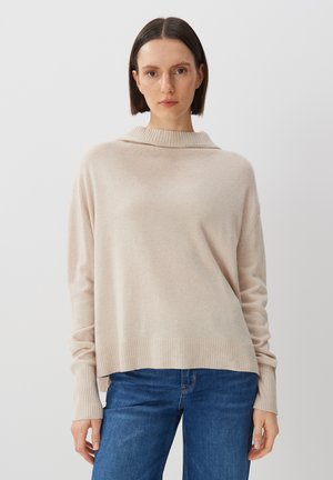 Beige Strickpullover mit hohem Kragen, lässiger Passform und gerippten Ärmelbündchen. Strukturierter Stoff und längerer Saum hinten, kombiniert mit blauen Jeans.