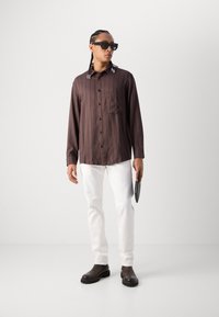 Chemise longue à manches longues à rayures verticales marron avec poche avant, pantalon blanc, bottines noires, et lunettes de soleil ; tenant une tablette noire.