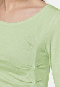 Hellgrünes langärmliges Oberteil mit einem runden Ausschnitt. Verfügt über dezente Rüschendetails an der Seite und einen weichen, strukturierten Stoff.