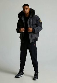 Černá prošívaná bunda s kapucí s umělým kožíškem, zapínáním na zip a nášivkou s logem. Černé kalhoty a šedé sportovní boty doplňují outfit.