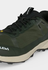 Παντόφλα πεζοπορίας σκούρου πράσινου και μαύρου χρώματος με μαύρα κορδόνια, ανθεκτική μπεζ σόλα και ορατό λογότυπο της μάρκας στα δάχτυλα και στη θηλιά της γλώσσας.