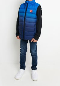 Modrý puff vest s přechodem od jasně modr�é po námořnickou, se středním zipem a kulatou nášivkou na hrudi. Přes černou košili s dlouhým rukávem.