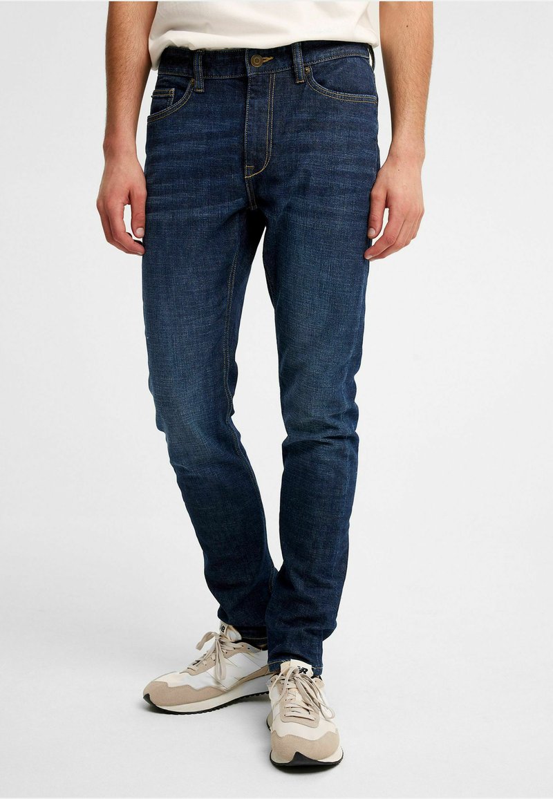 Mörkblå denimjeans med slim fit, försedd med guldtråd, klassisk femficksdesign och knappstängning. Bärs med ljusa sneakers.