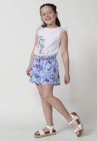 Camiseta blanca con mangas de malla abullonadas, que presenta un diseño gráfico. Pantalones cortos azules con estampado floral y una cinturilla elástica. Sandalias blancas.