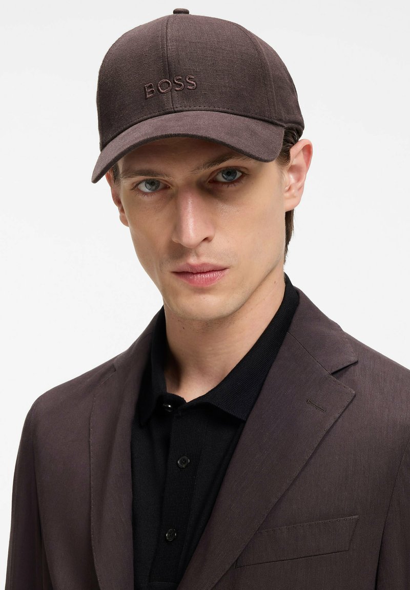 Homme portant une casquette marron foncé avec le logo "BOSS", un blazer marron assorti et une chemise noire à col, regardant droit devant lui.