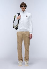 Vit zip-up jacka med hög krage, beige cargo-byxor med sidofickor och vita sneakers. Modell som bär solglasögon och bär en ryggsäck.