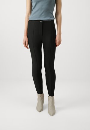 Leggings neri con vita alta, caratterizzati da cuciture frontali e una texture liscia, abbinati a stivaletti alla caviglia di colore chiaro.