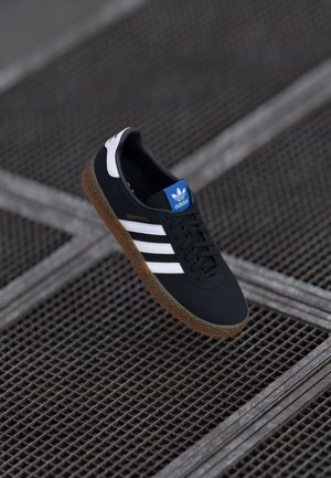 Czarny sneaker Adidas z białymi paskami, brązową podeszwą i niebieską etykietą na języku unosi się nad metalową kratką podłogową. Brak widocznych osób.