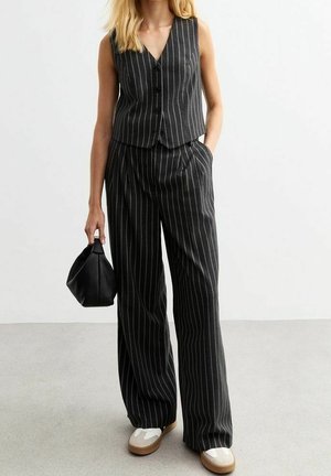 Kvinde iført sort stribet ærmeløs vest og matchende vidde bukser, holder en lille sort håndtaske, iført hvide og beige sko.