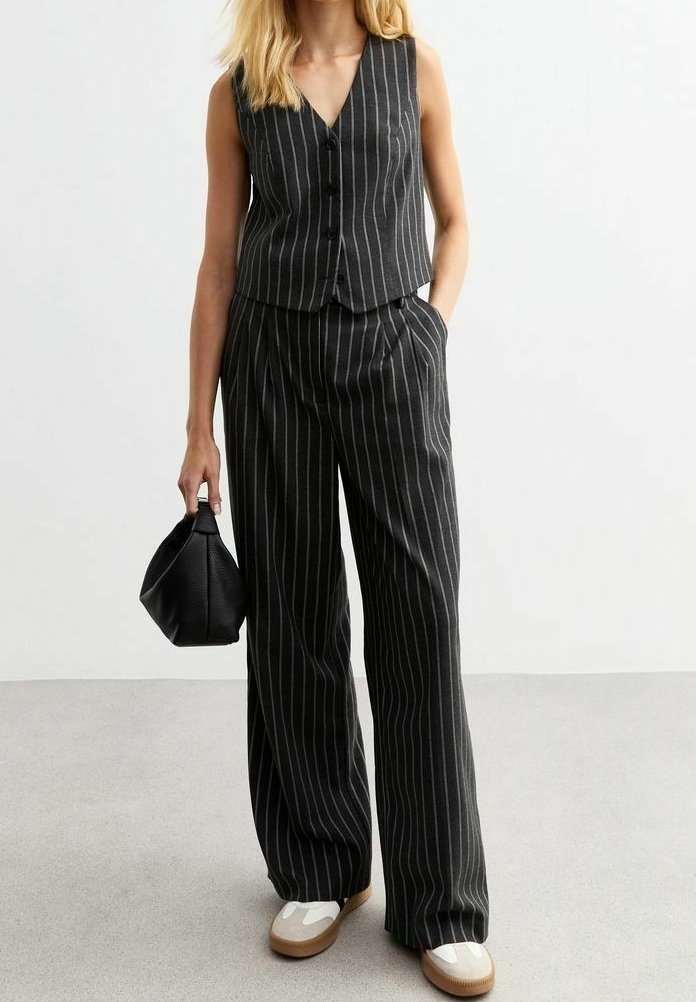 Femme portant un gilet sans manches à fines rayures noires et un pantalon large assorti, tenant un petit sac à main noir, chaussée de chaussures blanches et beiges.