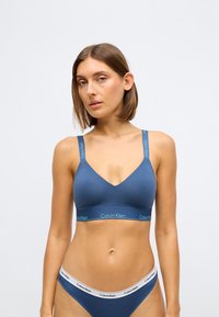 Bralette bleu Calvin Klein avec un design en V, des bretelles épaisses et une bande logo. Texture douce, style minimaliste, accompagné d'un bas de bikini assorti.