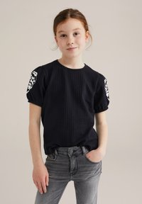 Crna kratka majica s kratkim rukavima od teksturiranog materijala, s bijelom cvjetnom vezom na rukavima, uparena sa sivim trapericama. Jednostavna okrugla izreza.