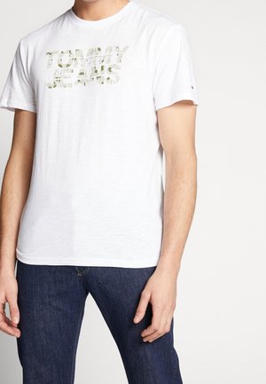 Mann trägt ein weißes kurzärmeliges T-Shirt mit "Tommy Jeans" in hellem Camouflage-Muster bedruckt, kombiniert mit dunkelblauen Jeans.